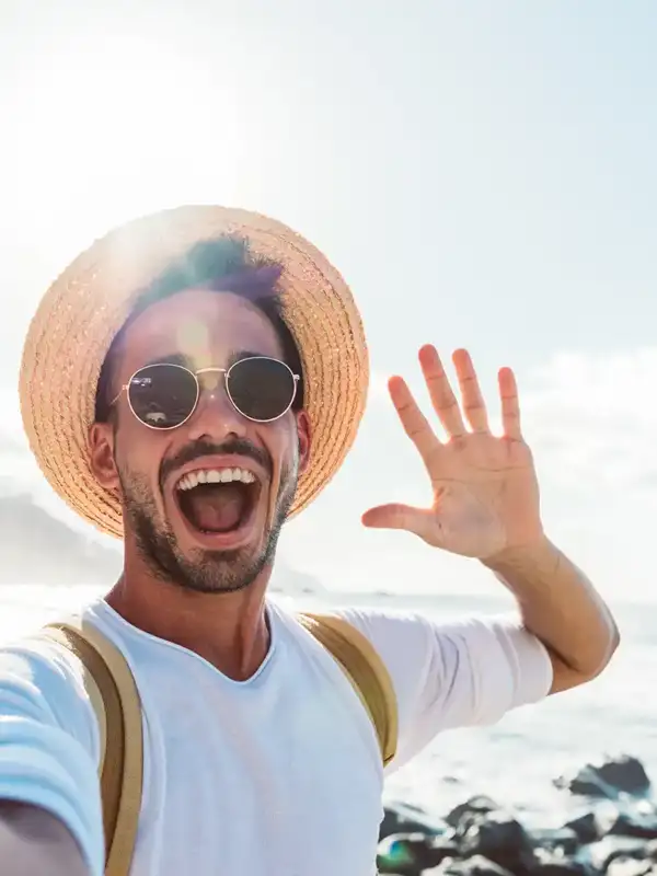 Selfie von einem männlichen Urlauber mit Sonnenhut der in die Kamera winkt.